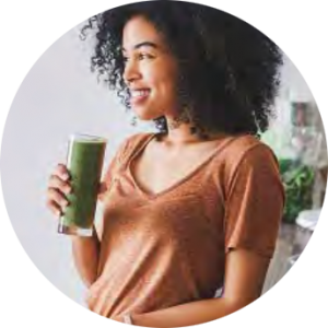 A smiling woman holding a green smoothie, indoors with soft light and casual attire.